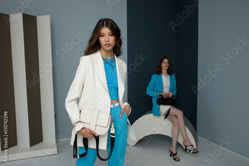 Two fashion models in aqua blue and white looks. Jacket, blazer, skirt, top, pants, trousers, handbags. Beautiful young women. Studio shot. Bicolor wall, light and dark blue. European and asian girls