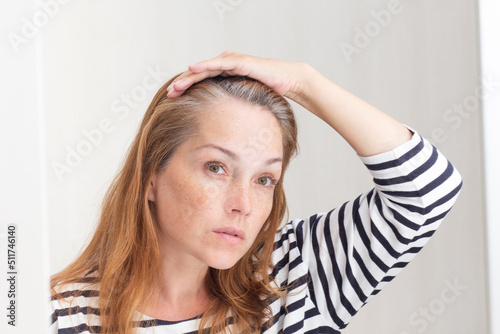 Serious 40s woman looking at gray hair in mirror reflection on growing out root