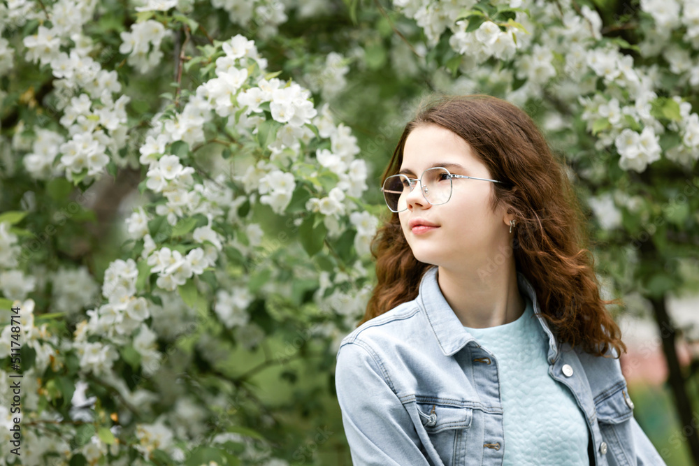 Portrait of beautiful caucasian teen girl with curly hair in glasses, looking confident, standing in blue jeans shirt on white blooming apple tree background outdoor. Portrait of beautiful caucasian teen girl with curly hair in glasses, looking confident, standing in blue jeans shirt on white blooming apple tree background outdoor.