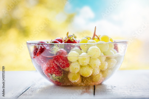 Transparent glass bowl filled with freshly picked strawberries and grapes, washed in water. Blurry bright sky and trees in the background. Summer organic farm fruits
