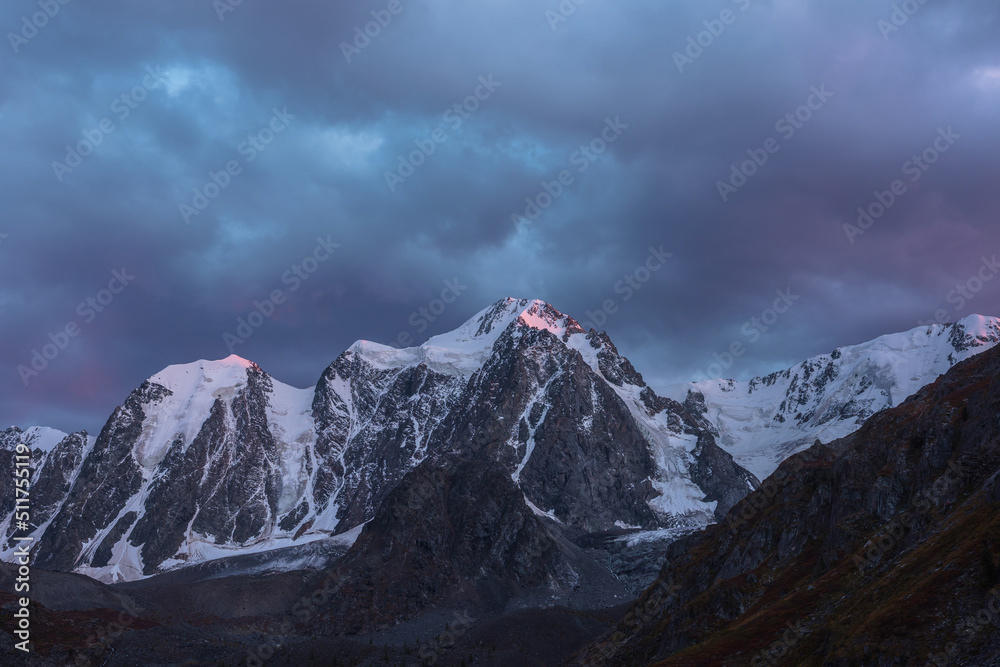 Awesome landscape with sunset pink reflection on huge snowy mountain top in violet dramatic sky. Hanging glacier and cornice on giant snow mountains in dusk. Snow-covered mountain range in twilight.