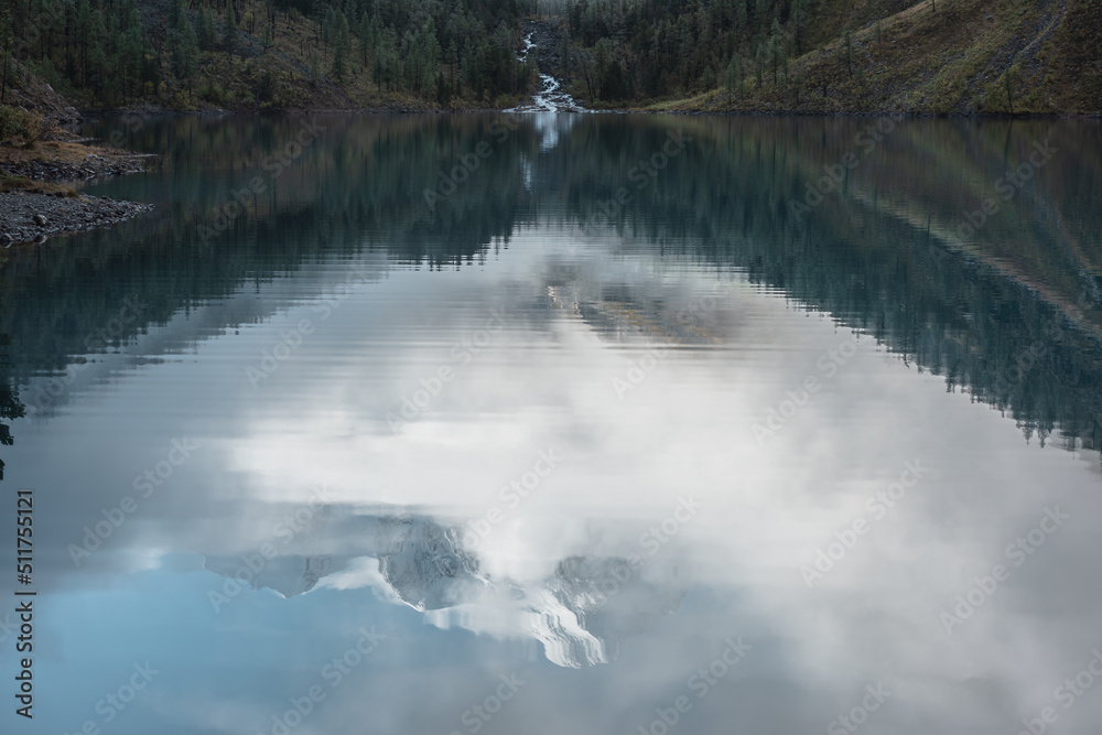 Tranquil scenery with reflex in mountain lake of snow castle in clouds. Snowy mountains in fog clearance, small river and coniferous trees reflected in alpine lake. Mountain creek and glacial lake.