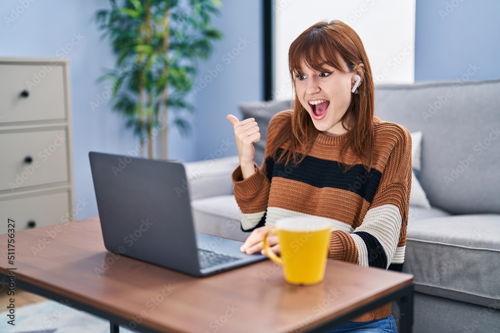 Young beautiful woman using computer laptop doing video call pointing thumb up to the side smiling happy with open mouth Young beautiful woman using computer laptop doing video call pointing thumb up to the side smiling happy with open mouth