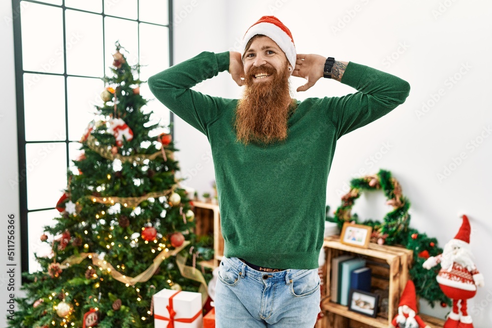 Redhead man with long beard wearing christmas hat by christmas tree relaxing and stretching, arms and hands behind head and neck smiling happy Redhead man with long beard wearing christmas hat by christmas tree relaxing and stretching, arms and hands behind head and neck smiling happy