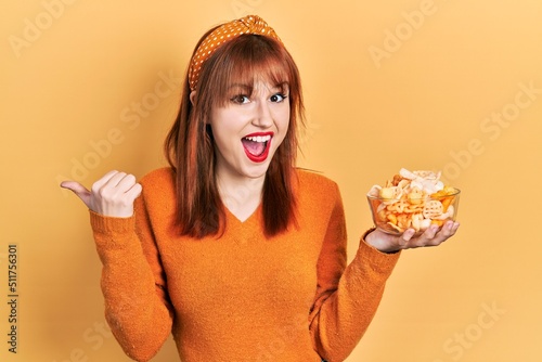 Redhead young woman holding potato chips pointing thumb up to the side smiling happy with open mouth