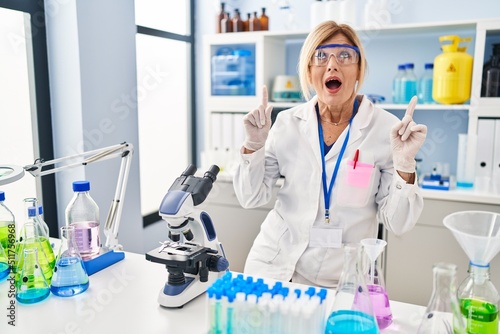 Middle age blonde woman working at scientist laboratory amazed and surprised looking up and pointing with fingers and raised arms.