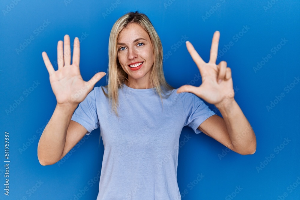 Fototapeta premium Beautiful blonde woman wearing casual t shirt over blue background showing and pointing up with fingers number eight while smiling confident and happy.