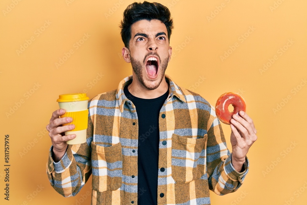 Fototapeta premium Young hispanic man eating doughnut and drinking coffee angry and mad screaming frustrated and furious, shouting with anger looking up.