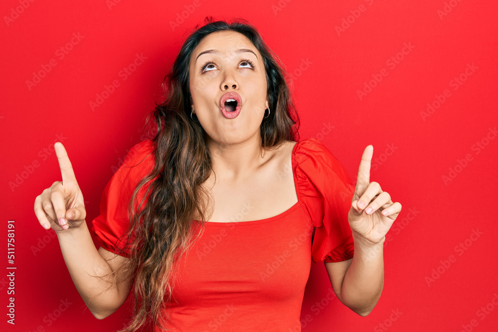 Young hispanic girl wearing casual clothes amazed and surprised looking up and pointing with fingers and raised arms. Young hispanic girl wearing casual clothes amazed and surprised looking up and pointing with fingers and raised arms.