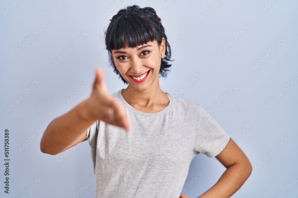 Young hispanic woman wearing casual t shirt over blue background smiling friendly offering handshake as greeting and welcoming. successful business. Young hispanic woman wearing casual t shirt over blue background smiling friendly offering handshake as greeting and welcoming. successful business.