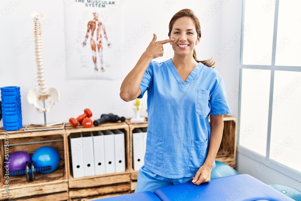 Middle age hispanic physiotherapist woman working at pain recovery clinic pointing with hand finger to face and nose, smiling cheerful. beauty concept Middle age hispanic physiotherapist woman working at pain recovery clinic pointing with hand finger to face and nose, smiling cheerful. beauty concept