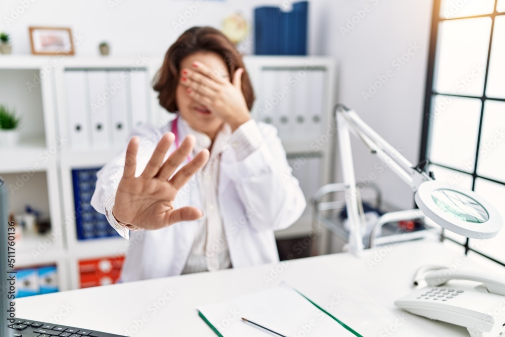 Middle age hispanic woman wearing doctor uniform and stethoscope at the clinic covering eyes with hands and doing stop gesture with sad and fear expression. embarrassed and negative concept. Middle age hispanic woman wearing doctor uniform and stethoscope at the clinic covering eyes with hands and doing stop gesture with sad and fear expression. embarrassed and negative concept.
