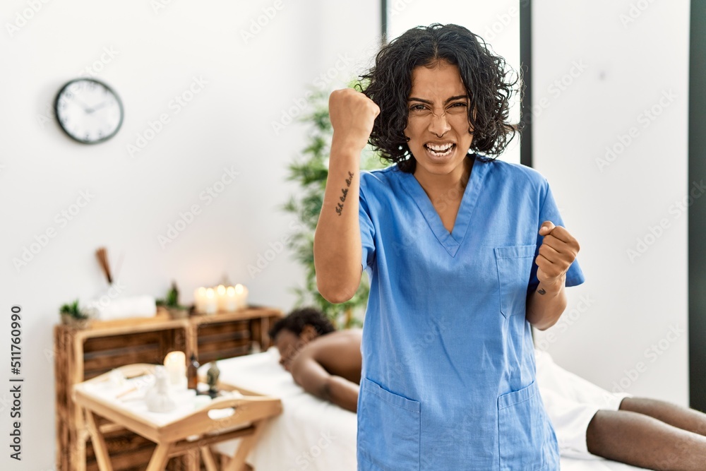 Young therapist woman at wellness spa center angry and mad raising fist frustrated and furious while shouting with anger. rage and aggressive concept. Young therapist woman at wellness spa center angry and mad raising fist frustrated and furious while shouting with anger. rage and aggressive concept.