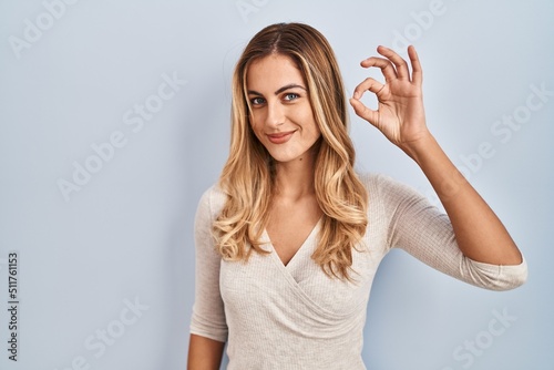 Young blonde woman standing over isolated background smiling positive doing ok sign with hand and fingers. successful expression.