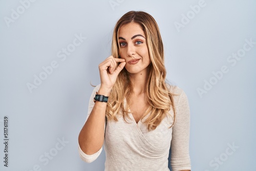 Young blonde woman standing over isolated background mouth and lips shut as zip with fingers. secret and silent, taboo talking