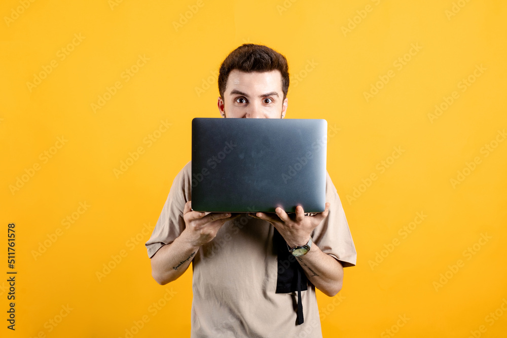 Handsome young man wearing casual clothes posing isolated over yellow background peeking out from behind laptop, looking surprised at camera.
