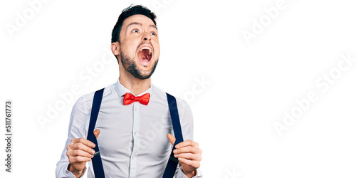 Hispanic man with beard wearing hipster look holding suspenders angry and mad screaming frustrated and furious, shouting with anger looking up.