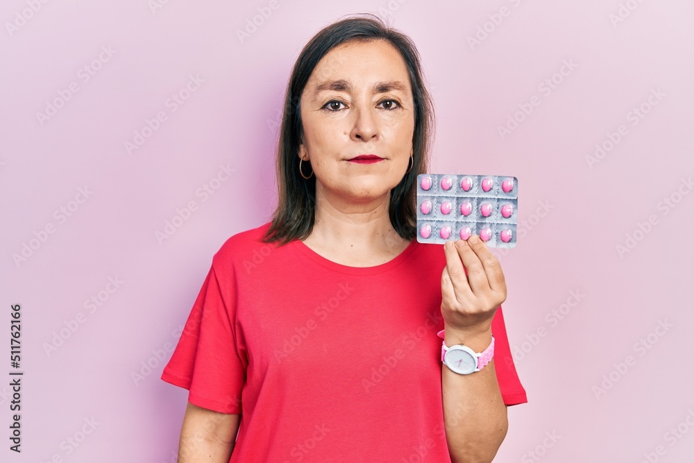 Middle age hispanic woman holding pills thinking attitude and sober expression looking self confident Middle age hispanic woman holding pills thinking attitude and sober expression looking self confident