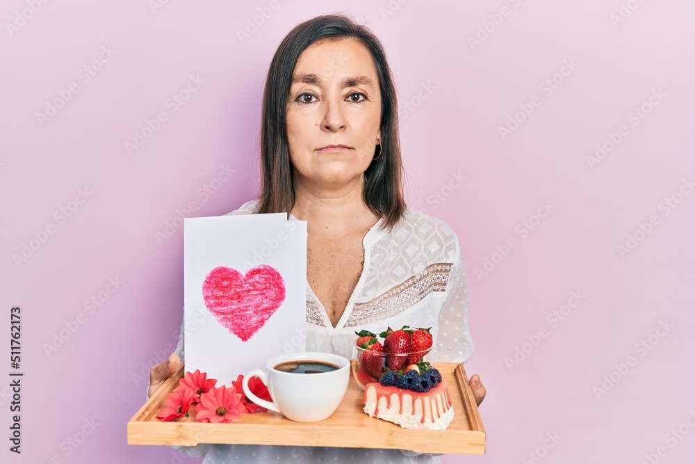 Middle age hispanic woman holding tray with breakfast food and heart draw relaxed with serious expression on face. simple and natural looking at the camera. Middle age hispanic woman holding tray with breakfast food and heart draw relaxed with serious expression on face. simple and natural looking at the camera.