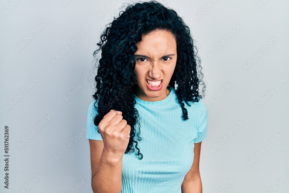 Young hispanic woman with curly hair wearing casual blue t shirt angry and mad raising fist frustrated and furious while shouting with anger. rage and aggressive concept.
