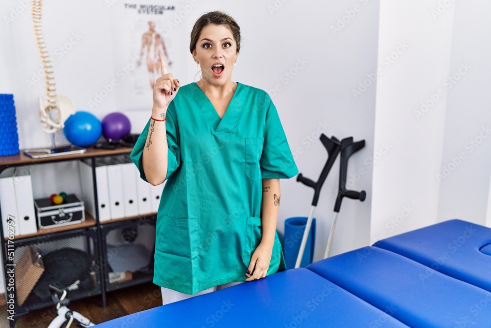 Young hispanic woman wearing physiotherapist uniform standing at clinic pointing finger up with successful idea. exited and happy. number one.