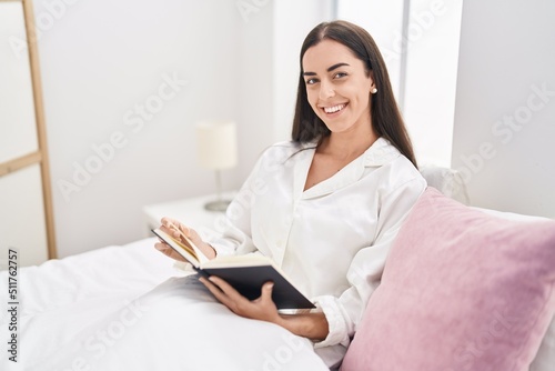 Young hispanic woman reading book sitting on bed at bedroom