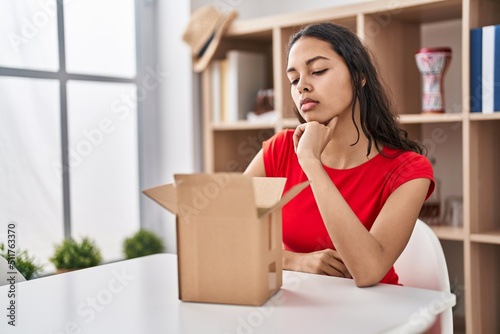 Young brazilian woman looking inside cardboard box thinking concentrated about doubt with finger on chin and looking up wondering