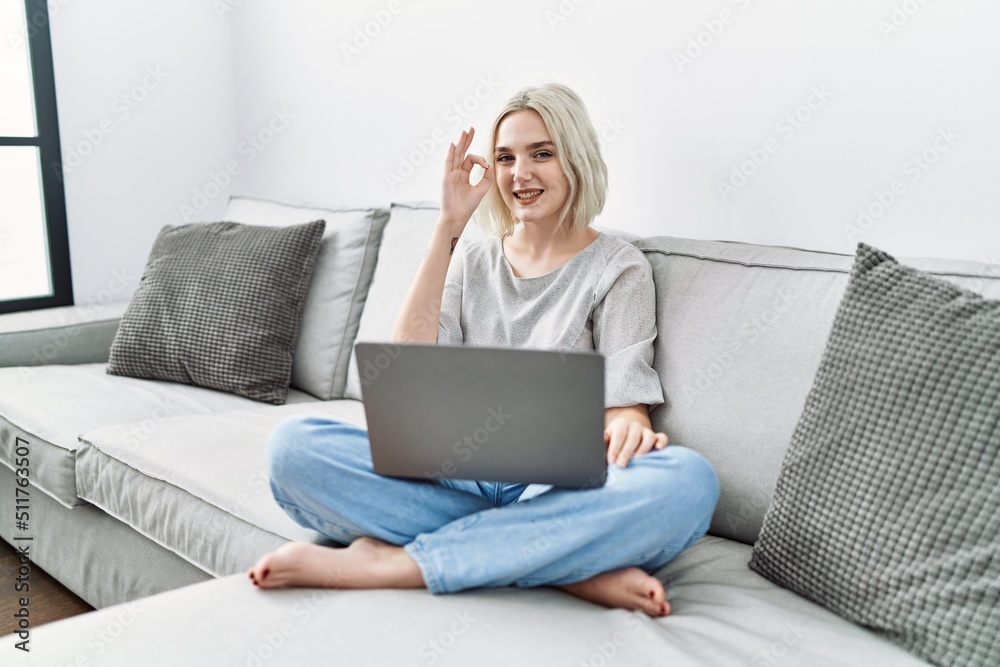 Young caucasian woman using laptop at home sitting on the sofa smiling positive doing ok sign with hand and fingers. successful expression. Young caucasian woman using laptop at home sitting on the sofa smiling positive doing ok sign with hand and fingers. successful expression.