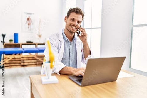 Young hispanic man wearing physiotherapist uniform using laptop talking on the smartphone at clinic