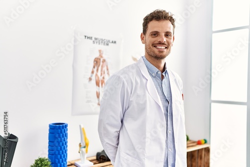 Young hispanic man wearing physiotherapist uniform standing at clinic