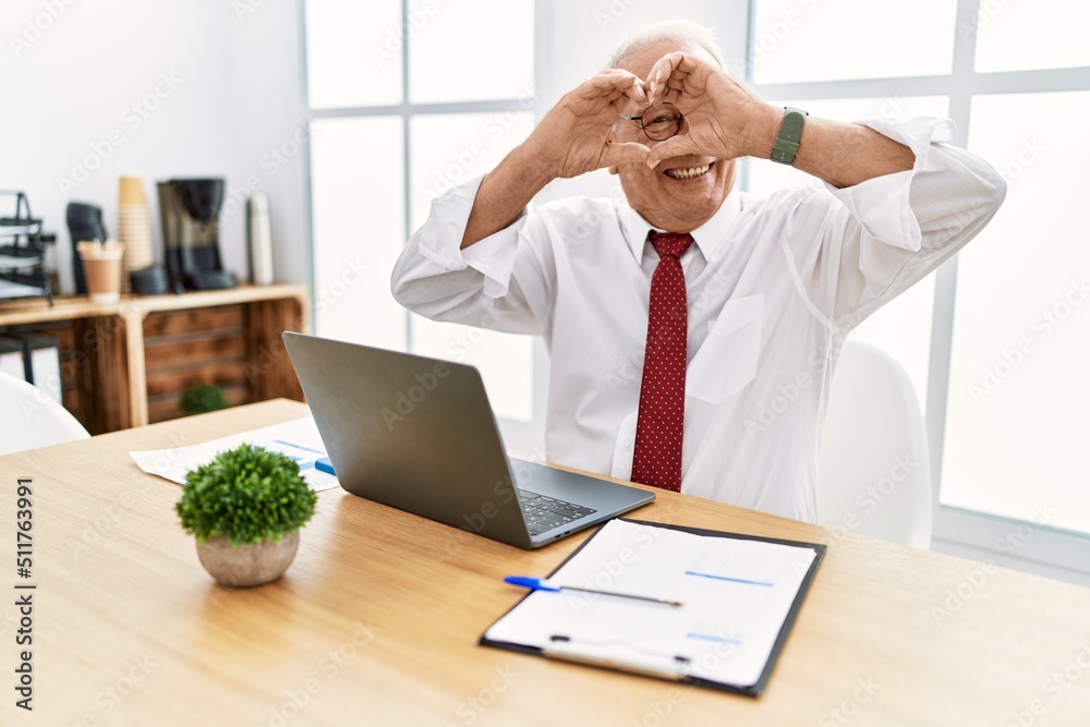 Senior man working at the office using computer laptop doing heart shape with hand and fingers smiling looking through sign Senior man working at the office using computer laptop doing heart shape with hand and fingers smiling looking through sign