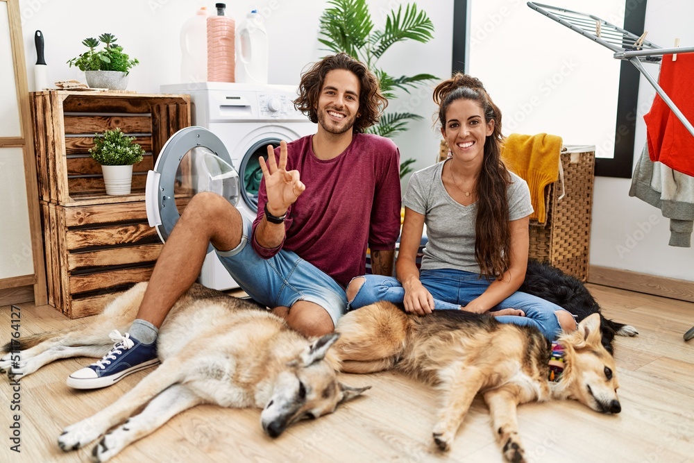 Young hispanic couple doing laundry with dogs showing and pointing up with fingers number three while smiling confident and happy.