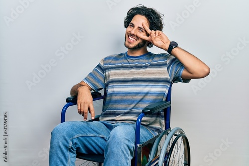 Handsome hispanic man sitting on wheelchair doing peace symbol with fingers over face, smiling cheerful showing victory