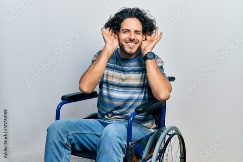 Handsome hispanic man sitting on wheelchair trying to hear both hands on ear gesture, curious for gossip. hearing problem, deaf