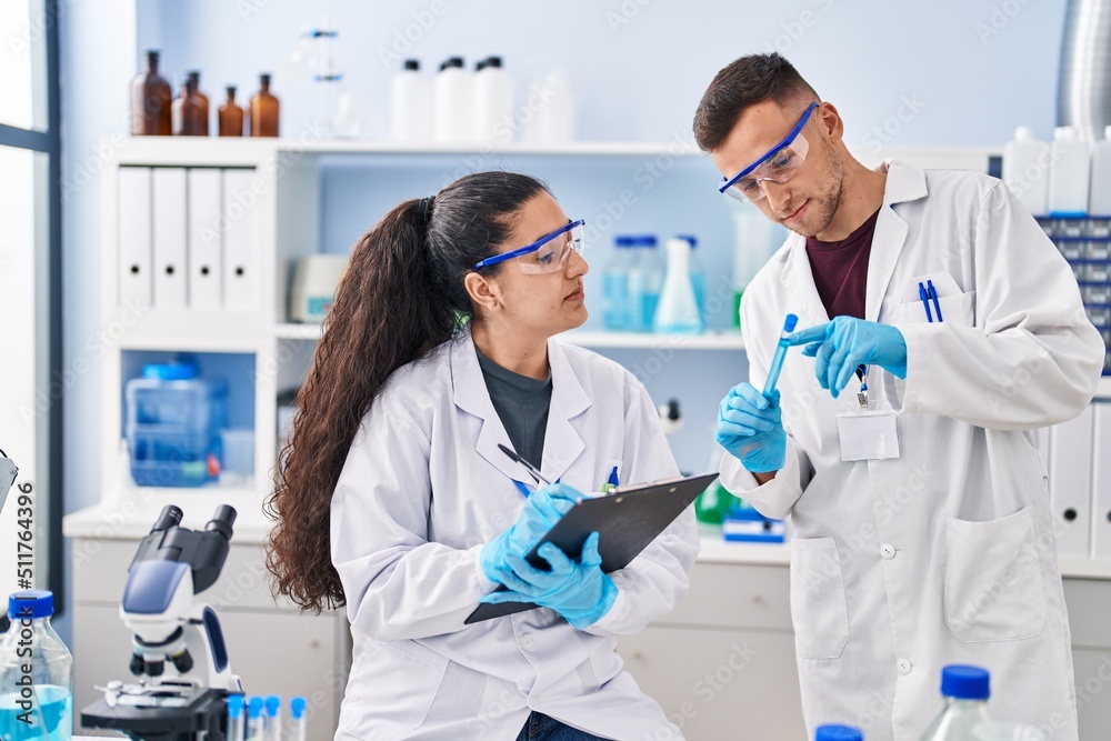 Man and woman wearing scientist uniform write on clipboard holding test tube at laboratory Man and woman wearing scientist uniform write on clipboard holding test tube at laboratory