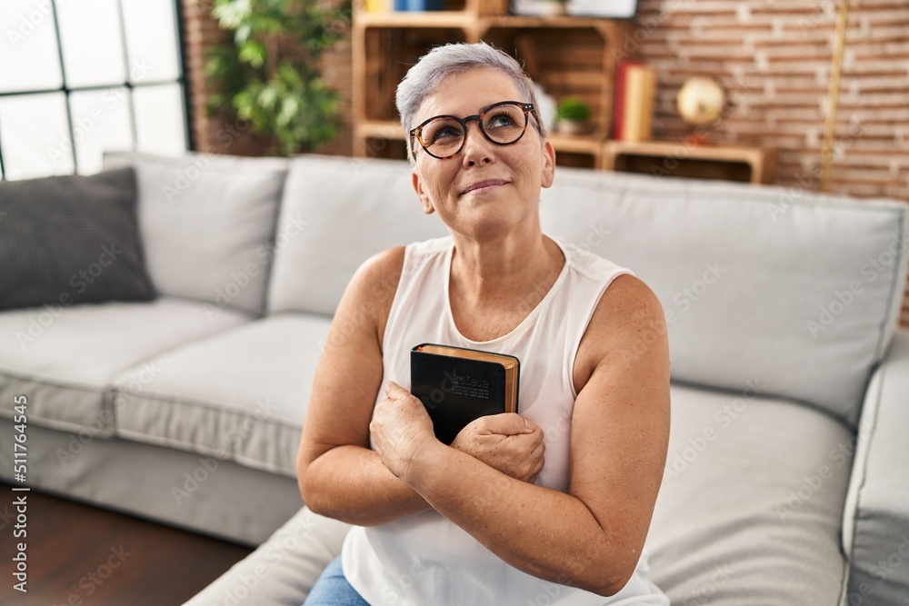 Middle age woman hugging bible sitting on sofa at home Middle age woman hugging bible sitting on sofa at home