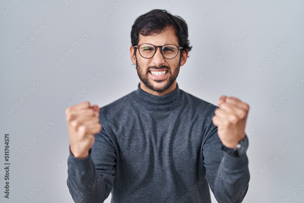 Handsome latin man standing over isolated background angry and mad raising fists frustrated and furious while shouting with anger. rage and aggressive concept. Handsome latin man standing over isolated background angry and mad raising fists frustrated and furious while shouting with anger. rage and aggressive concept.