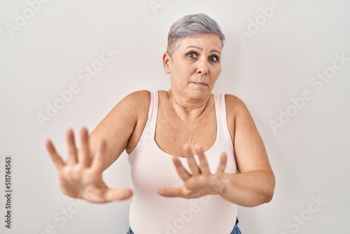 Middle age caucasian woman standing over white background afraid and terrified with fear expression stop gesture with hands, shouting in shock. panic concept.