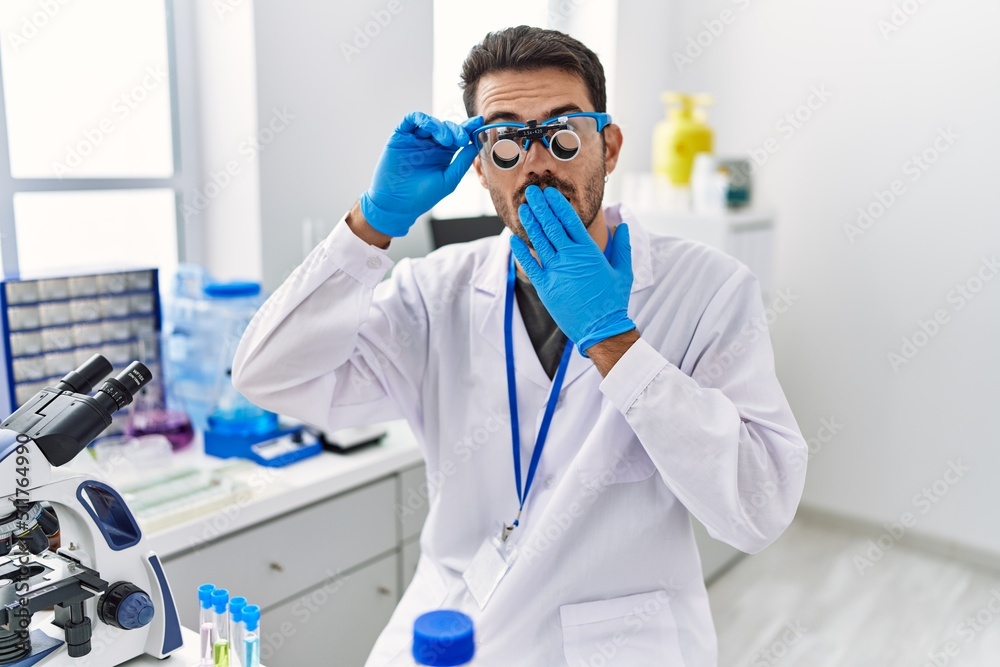 Young hispanic man working at scientist laboratory wearing magnifying glasses covering mouth with hand, shocked and afraid for mistake. surprised expression Young hispanic man working at scientist laboratory wearing magnifying glasses covering mouth with hand, shocked and afraid for mistake. surprised expression