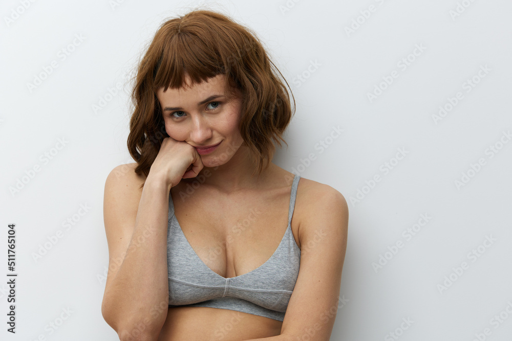 a funny, slightly upset tanned woman with red hair stands on a light background in a gray top and supports her cheek with a fist of one hand a funny, slightly upset tanned woman with red hair stands on a light background in a gray top and supports her cheek with a fist of one hand