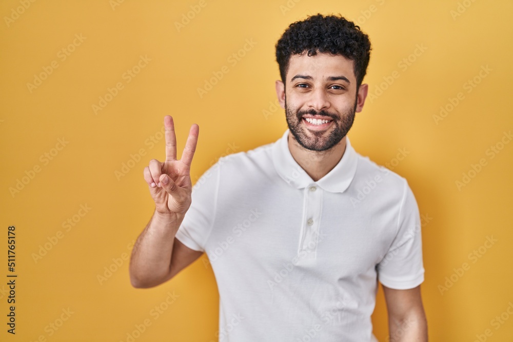 Arab man standing over yellow background smiling looking to the camera showing fingers doing victory sign. number two. Arab man standing over yellow background smiling looking to the camera showing fingers doing victory sign. number two.