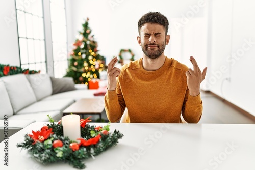 Arab young man sitting on the table by christmas tree gesturing finger crossed smiling with hope and eyes closed. luck and superstitious concept.