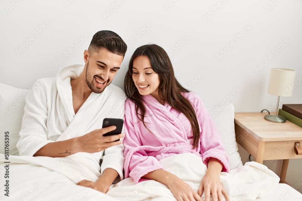 Young hispanic couple using smartphone lying on bed at home.