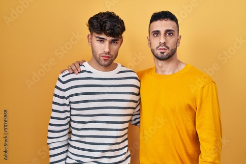 Young hispanic gay couple standing over yellow background relaxed with serious expression on face. simple and natural looking at the camera.