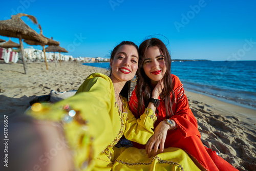 Two Arab women, photo taken in selfie mode on the beach sitting with typical Moroccan clothes. Enjoying the summer, having a great time. Horizontal photo