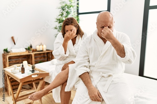 Middle age hispanic couple wearing bathrobe at wellness spa tired rubbing nose and eyes feeling fatigue and headache. stress and frustration concept.