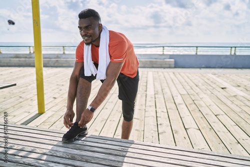 Fototapeta Naklejka Na Ścianę i Meble -  Young african american man wearing sportswear tying shoe at seaside