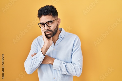 Hispanic man with beard standing over yellow background thinking looking tired and bored with depression problems with crossed arms.