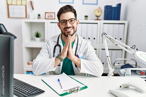 Young man with beard wearing doctor uniform and stethoscope at the clinic praying with hands together asking for forgiveness smiling confident.