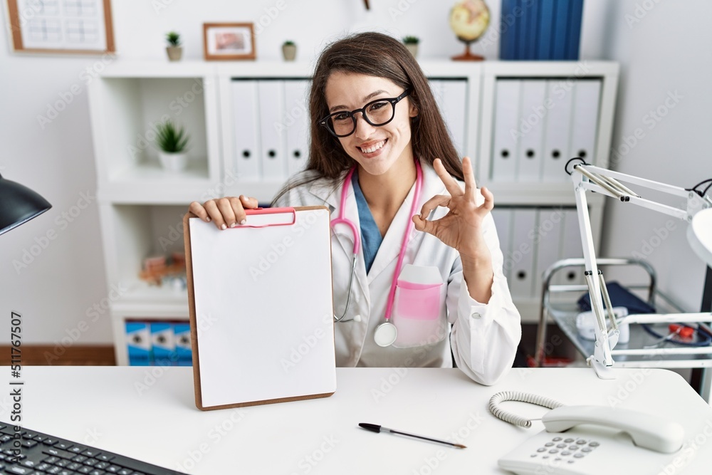Young doctor woman holding clipboard at the clinic doing ok sign with fingers, smiling friendly gesturing excellent symbol Young doctor woman holding clipboard at the clinic doing ok sign with fingers, smiling friendly gesturing excellent symbol